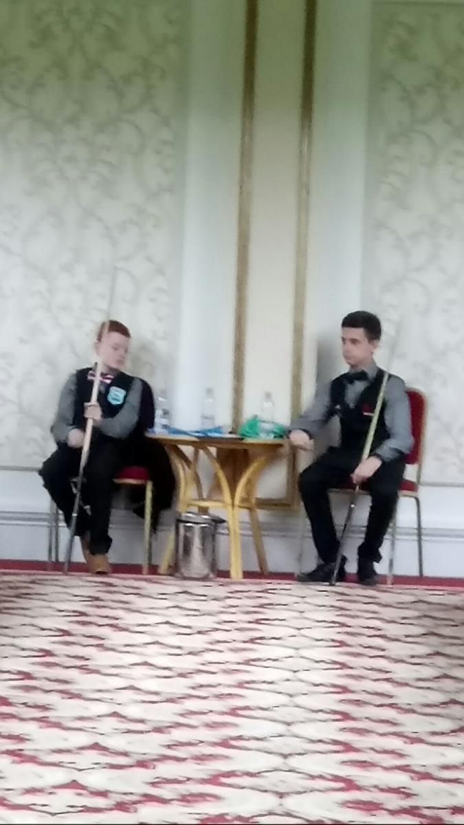 Twee mannen in vesten en vlinderdassen zitten met keuen in de hand aan een tafel. Ze bevinden zich in een kamer met sierlijke decoraties.