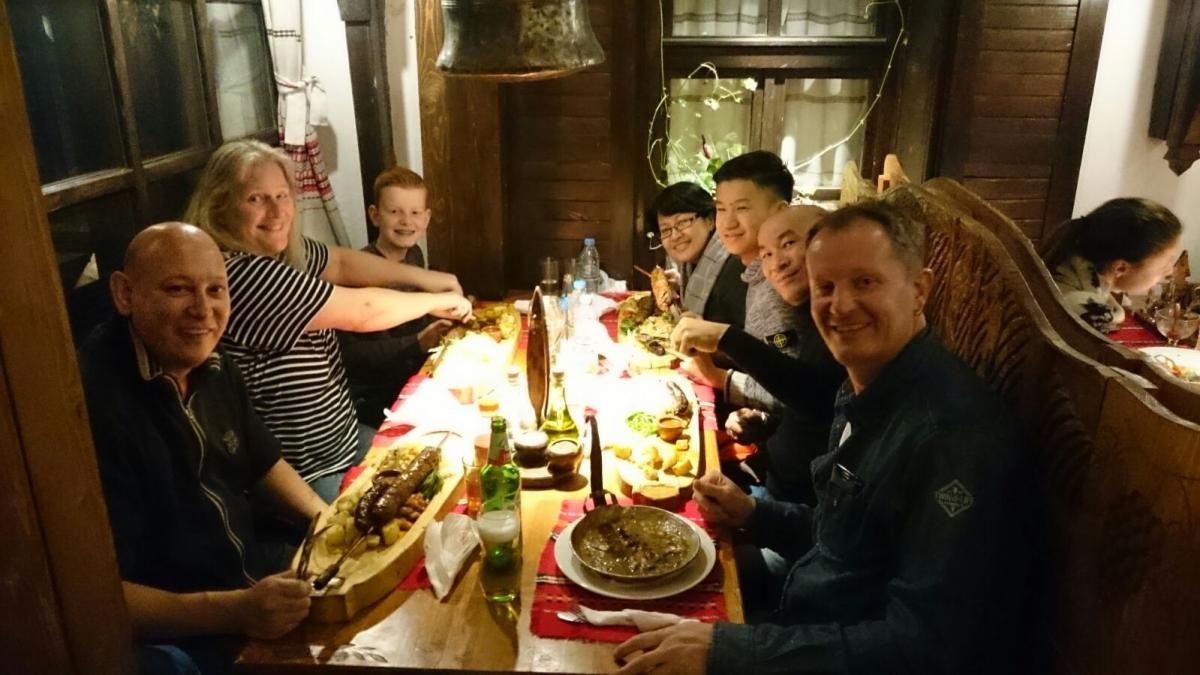 Een groep mensen eet aan een houten tafel, met dienbladen in de handen, in een rustiek, warm verlicht restaurant.
