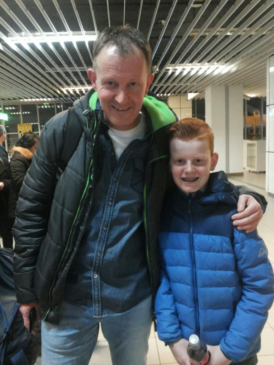 Man met arm om een ​​jonge jongen, beiden lachend. Ze zijn binnen, de jongen heeft rood haar en een blauwe jas.