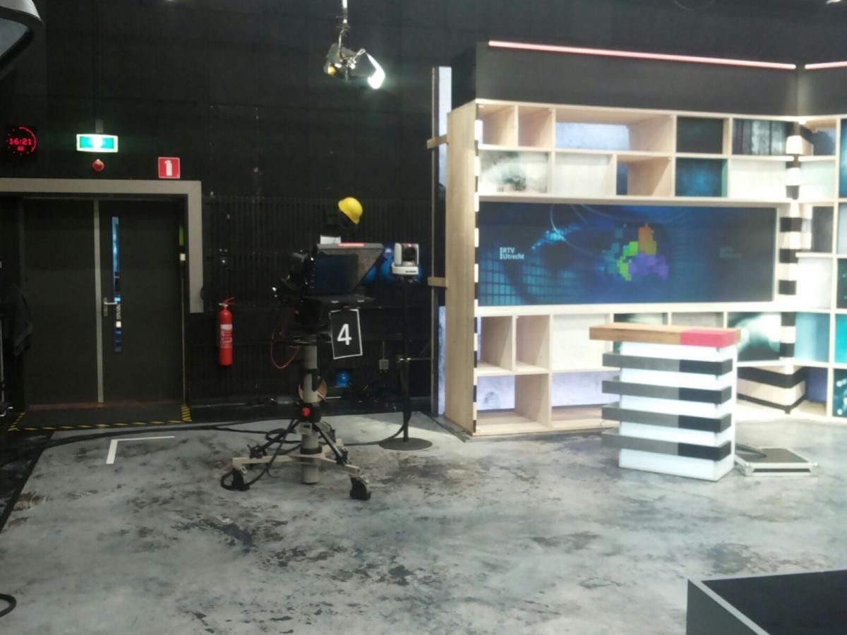 Televisiestudio met camera op statief, opstelling met digitaal scherm en stellingen.