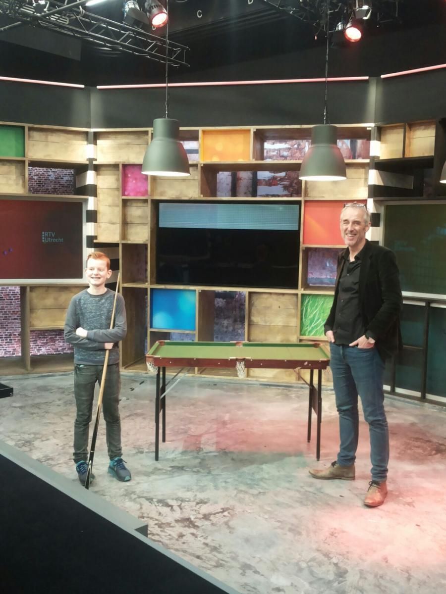 Een jongen en een man staan ​​bij een tafel in een kleurrijke tv-studio. De jongen houdt een keu vast en de man glimlacht.