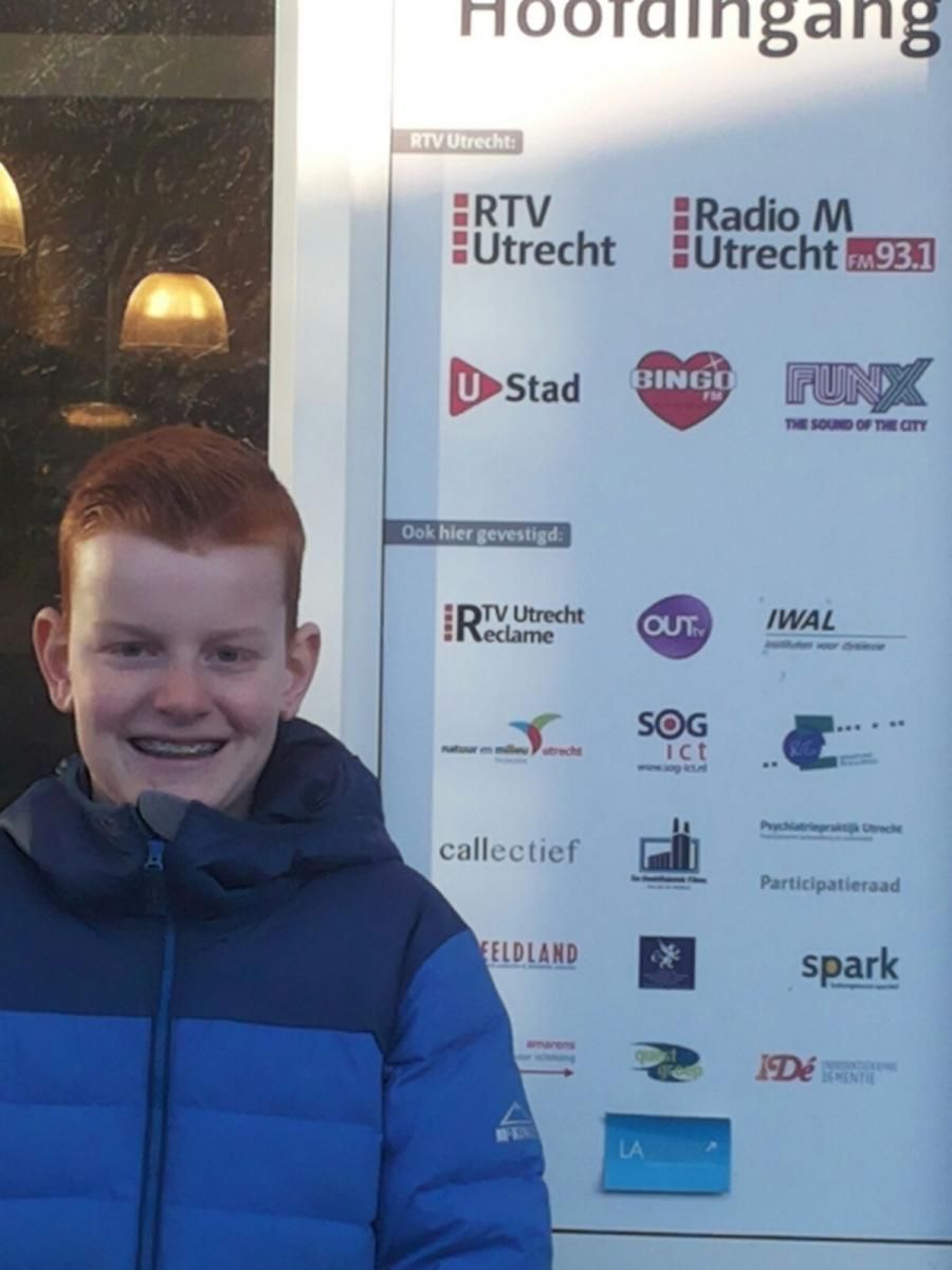 Een jonge jongen met rood haar lacht voor een bord met logo's van RTV Utrecht en andere organisaties.