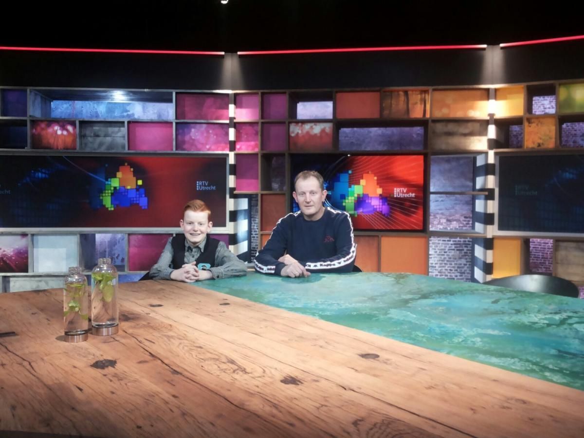 Twee mensen zitten aan een tafel in een kleurrijke studio. Een van hen is een kind. Achter hen een muur van schermen.