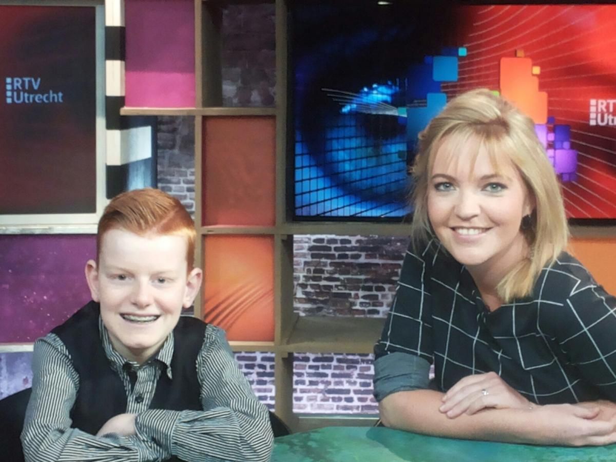 Een jonge jongen en een vrouw lachen naar de camera in een kleurrijke tv-studio. De logo's van 