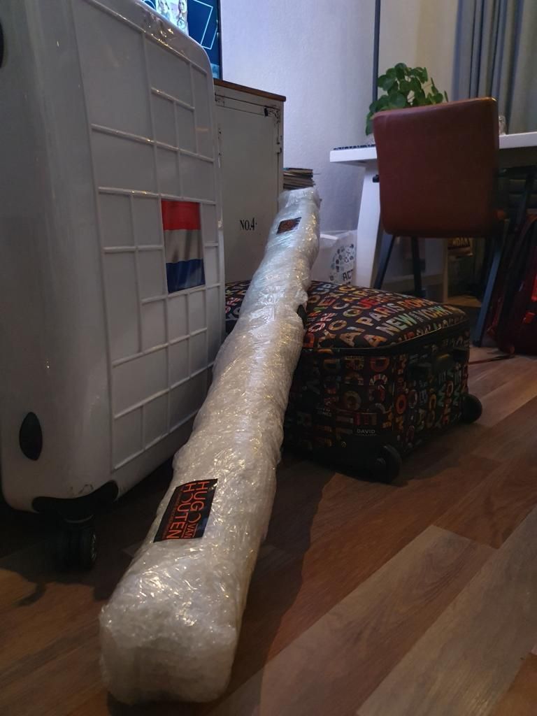 Een lang, in bubbeltjesplastic verpakt pakket staat op de houten vloer naast een witte kast en een voetenbankje met patroon.