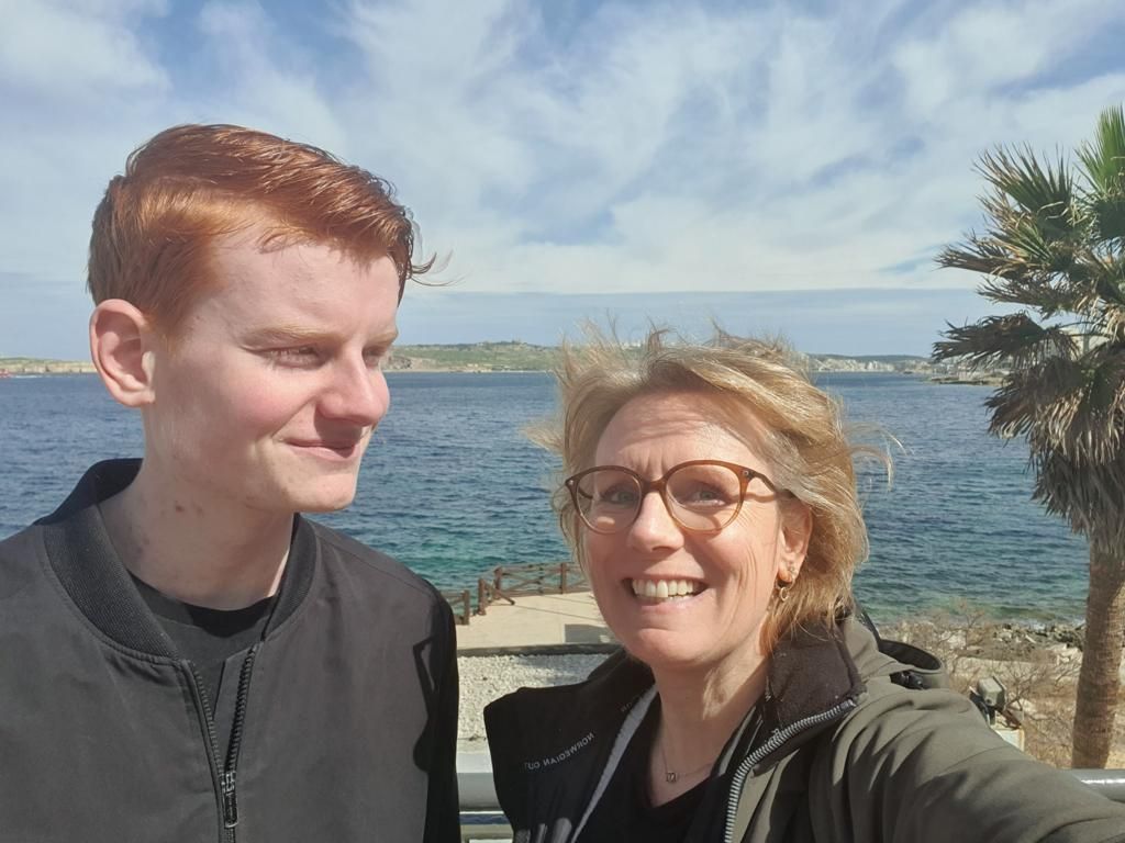 Jonge man en oudere vrouw lachen voor een selfie bij de oceaan, met palmbomen en blauwe lucht.