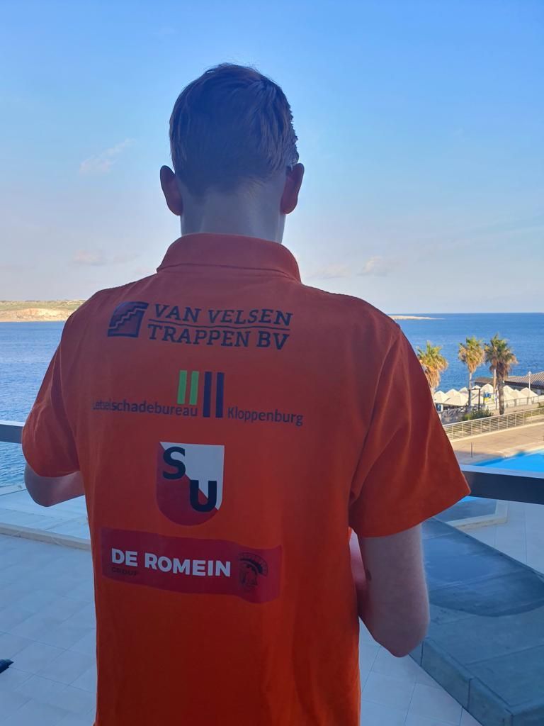 Persoon in oranje shirt met logo's, uitkijkend over zee.