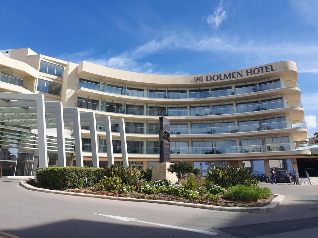 Het Dolmen Hotelgebouw op Malta met een gebogen gevel, een blauwe lucht en groen op de voorgrond.