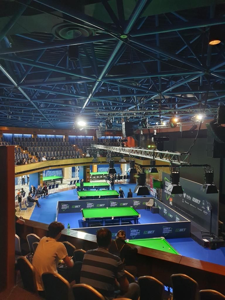 Snookertoernooizaal met meerdere tafels. Toeschouwers kijken toe vanaf tribunes. Blauwe en groene tinten domineren.