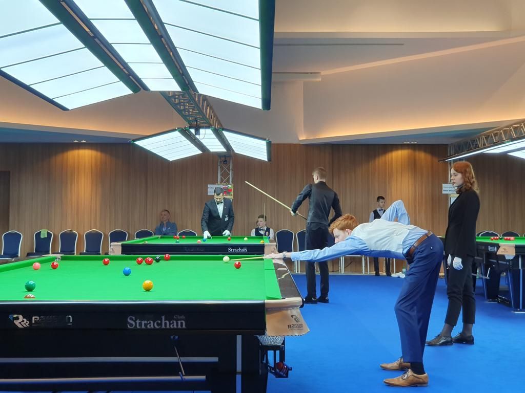 Snookerwedstrijd: speler richt keu. Groene tafel, blauwe vloer. Publiek kijkt toe. Lichte kamer.