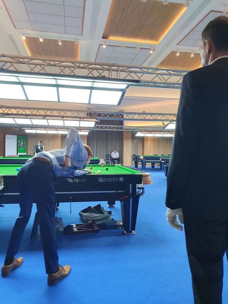 Snookerspelers in actie aan een groene tafel in een felverlichte overdekte arena; blauwe vloer, scheidsrechter.