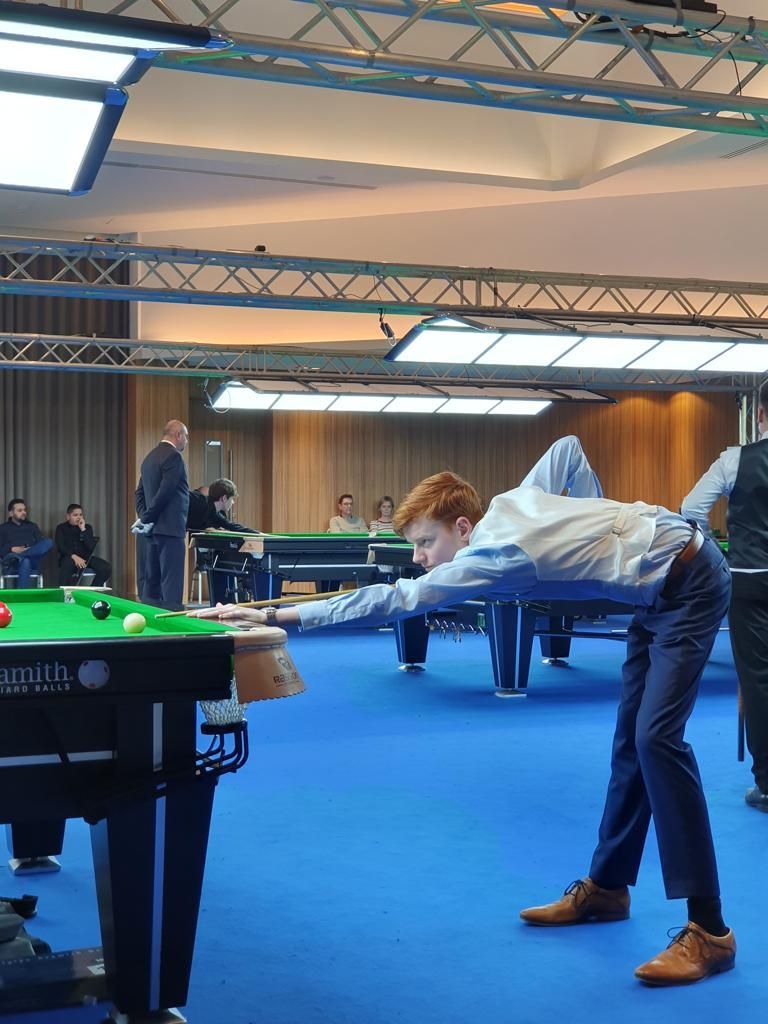 Een jongeman met rood haar in een formeel pak speelt snooker en richt op de speelbal op een blauwe vilten tafel.