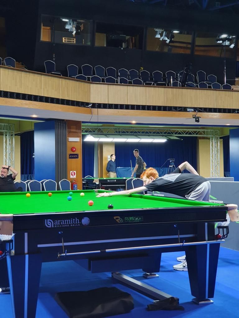 Snookerspeler in een stadion, die een bal probeert te slaan. Groene tafel, blauwe rand, spelers en toeschouwers aanwezig.
