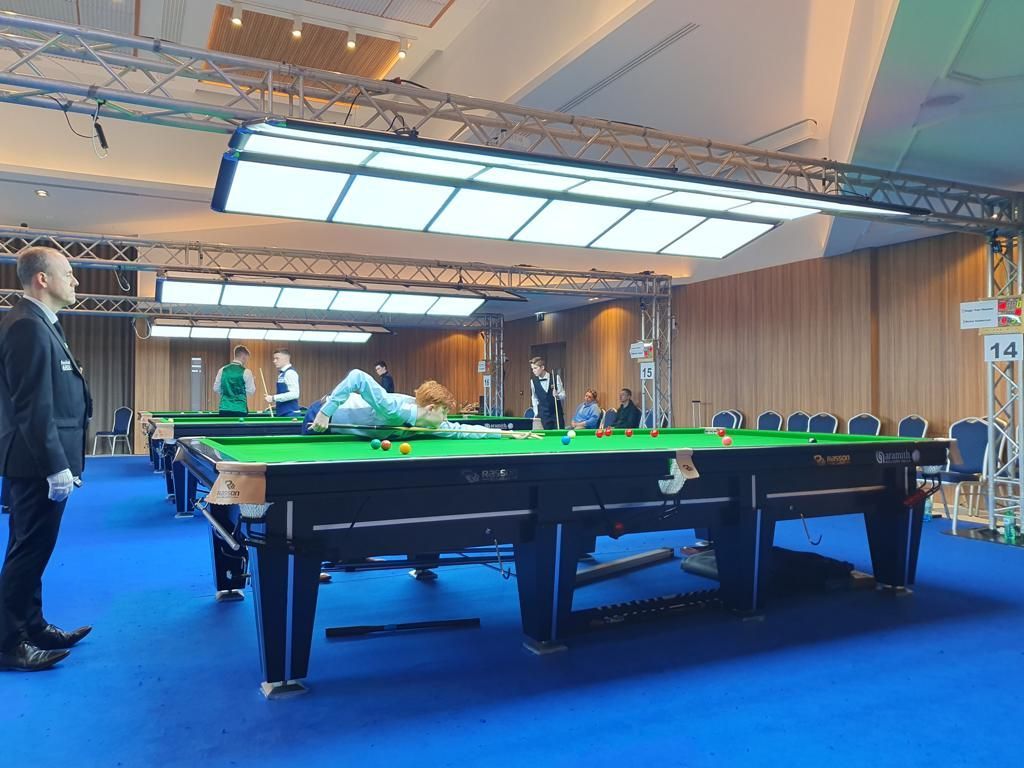 Snookertoernooi in een grote zaal, spelers aan groene tafels, scheidsrechters in zwarte pakken en publiek.