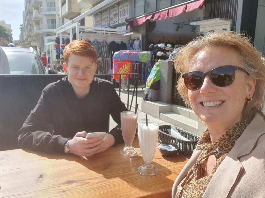 Vrouw en roodharige tiener lachend aan een tafeltje met drankjes buiten. Zonnige dag; gebouwen en winkels op de achtergrond.