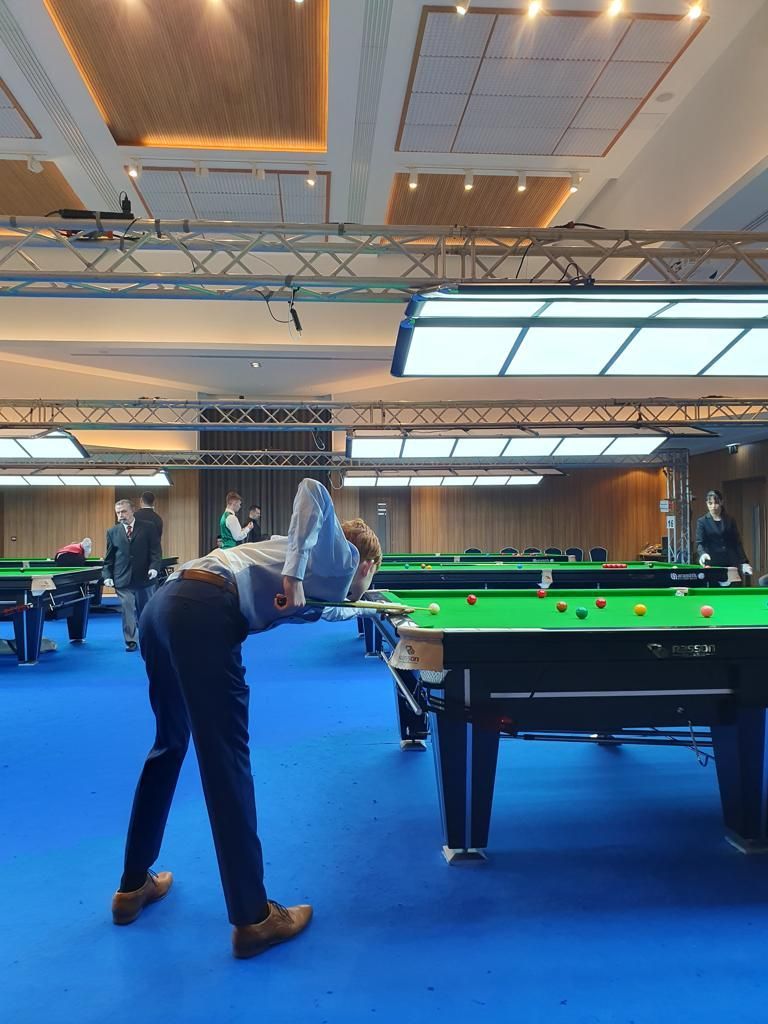 Een man leunt over een snookertafel in een hal met een blauwe vloer en maakt zich klaar om een ​​balletje te slaan.