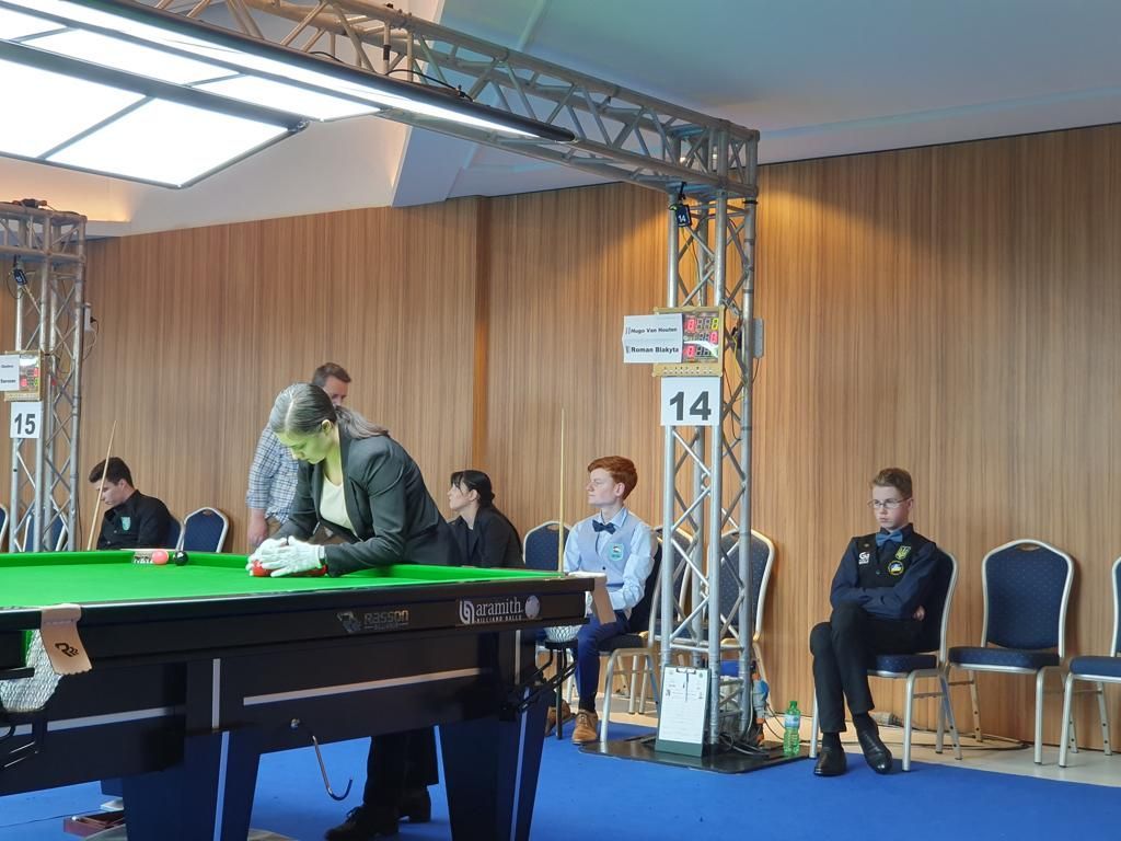 Snookerwedstrijd. Een speler in een grijs pak leunt over een tafel. Anderen kijken toe, waaronder een jongen met een vlinderdas. Binnen in een wedstrijdlocatie.