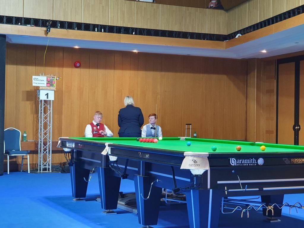 Snookerwedstrijd: spelers en scheidsrechter aan een groene tafel met gekleurde ballen, binnen.