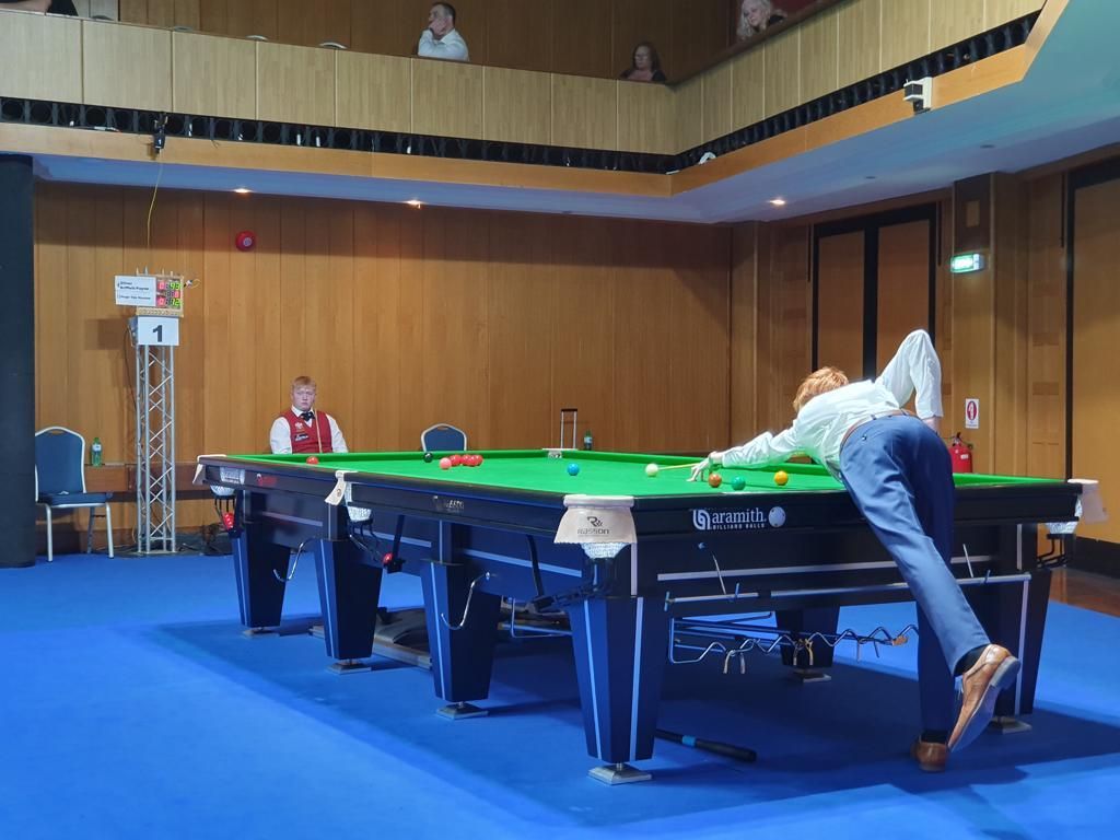 Een persoon speelt snooker en mikt op ballen op een groene tafel. Een ander kijkt toe. Blauw tapijt, houten lambrisering.