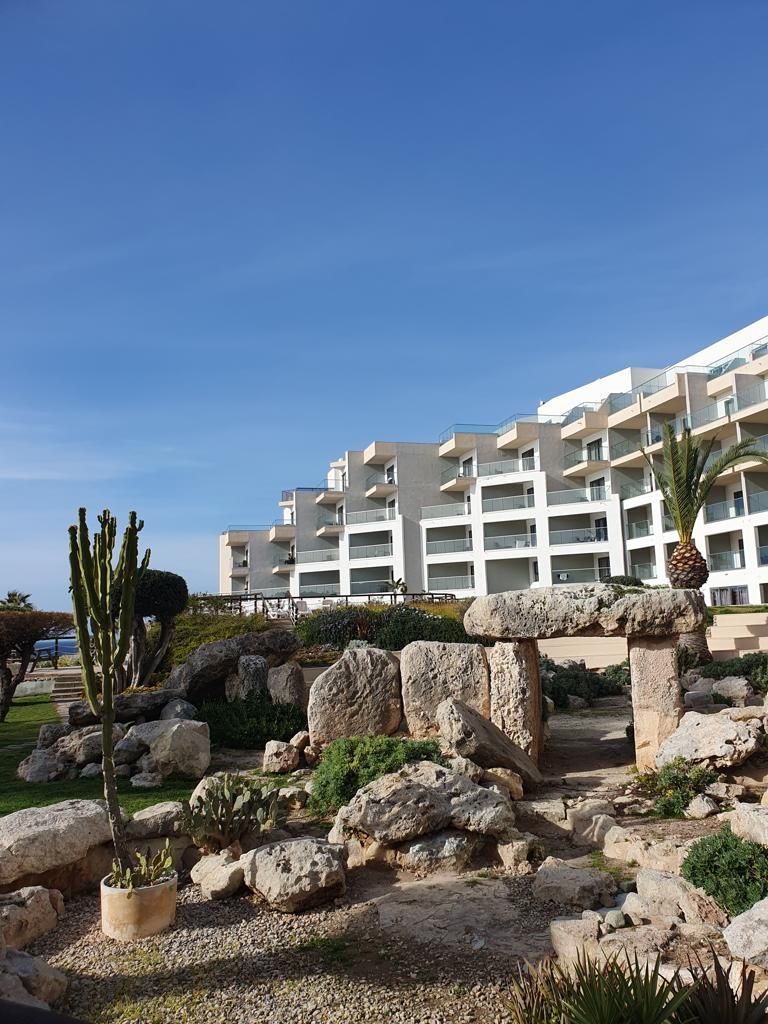 Oude stenen bouwwerken in een tuin met op de achtergrond een groot, wit hotel onder een blauwe lucht.