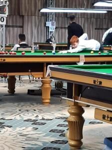 Snookerwedstrijd: Een speler in het wit leunt over een groene tafel en mikt. Andere spelers op de achtergrond.