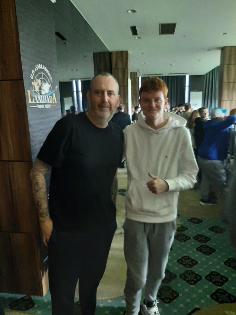 Twee mannen poseren samen binnen. De een met tatoeages en een zwart shirt, de ander een roodharige in een witte hoodie die een duim omhoog steekt.