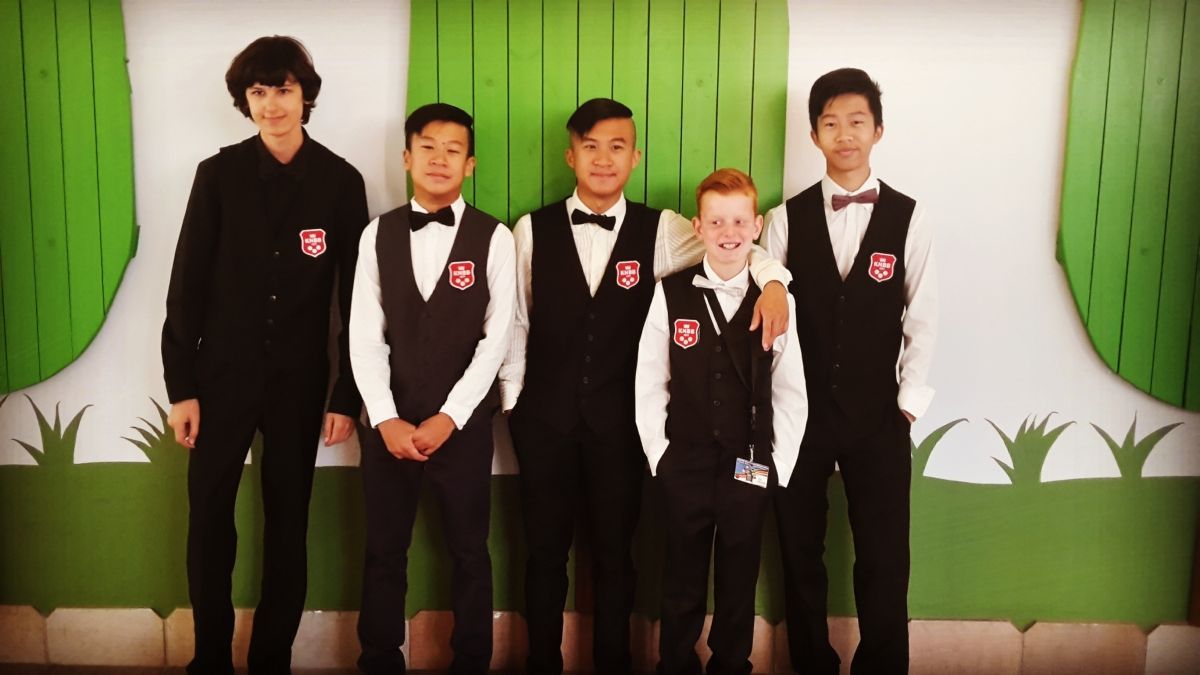 Vijf jongens in zwarte vesten en vlinderdassen poseren voor een groen-witte achtergrond.