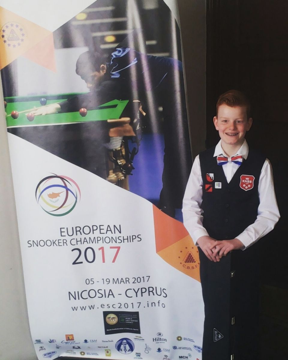 Een jonge jongen in een snookeruniform poseert naast een spandoek met reclame voor de Europese Snookerkampioenschappen van 2017 in Cyprus.