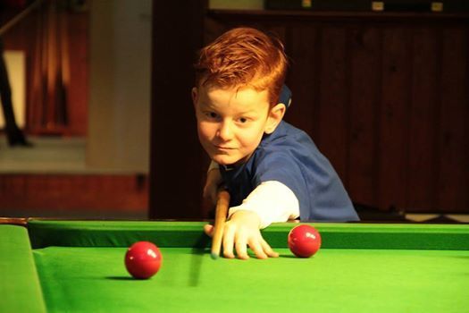 Een jongen met rood haar richt een keu op rode ballen op een groene tafel in een slecht verlichte kamer.