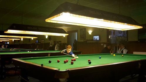 Een persoon speelt snooker in een schemerige zaal met meerdere tafels onder rechthoekige lampen.