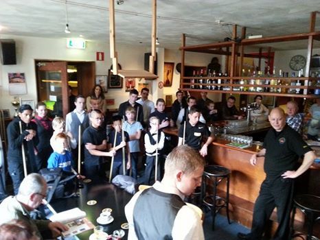 Mensen verzamelden zich in een bar, velen met poolkeuen in de hand. Een groep kinderen en volwassenen is aanwezig bij de bar.