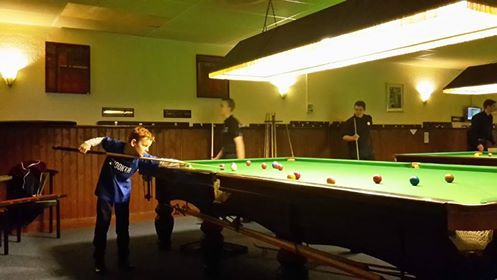 Een jongen staat klaar om een ​​stoot te spelen aan een snookertafel. Op de achtergrond zijn andere spelers te zien bij een snookerclub.