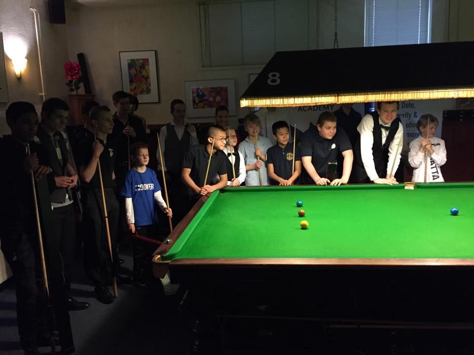 Een groep mensen kijkt naar een snookerwedstrijd. Spelers en toeschouwers staan ​​binnen rond de tafel.