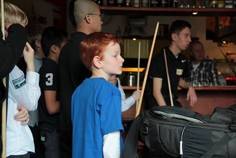 Een jongen met rood haar in een blauw shirt kijkt in een poolzaal naar een wedstrijd. Andere spelers en een bar op de achtergrond.