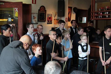 Een groep mensen, sommigen met keuen in de hand, kijkt in een bar naar een poolwedstrijd.
