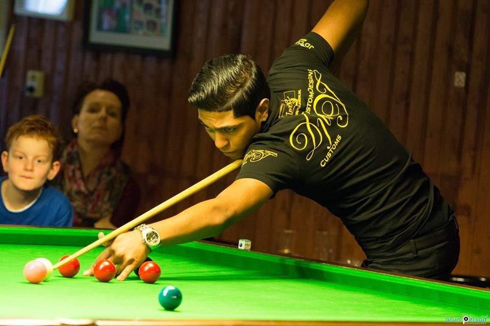 Een man in een zwart shirt speelt snooker en richt op rode ballen. Op de achtergrond kijken twee mensen toe.