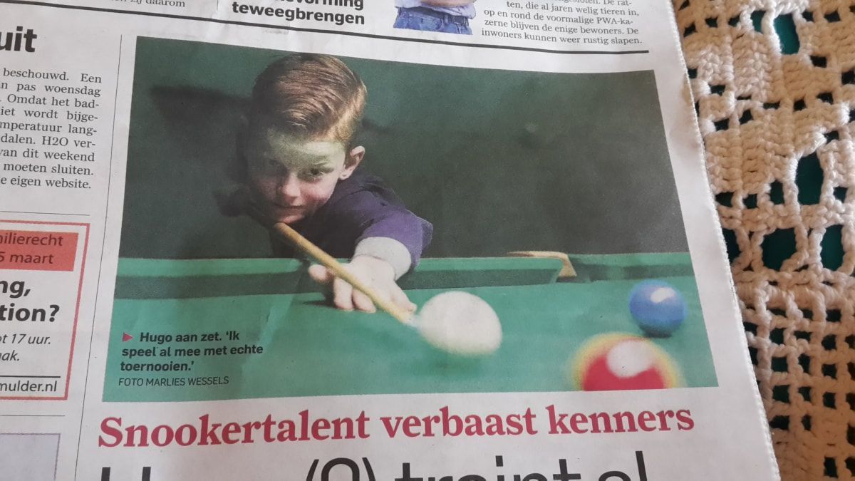 Een jongen speelt snooker en richt zijn keu op de witte bal op een groene tafel. Rode en blauwe ballen liggen vlakbij.