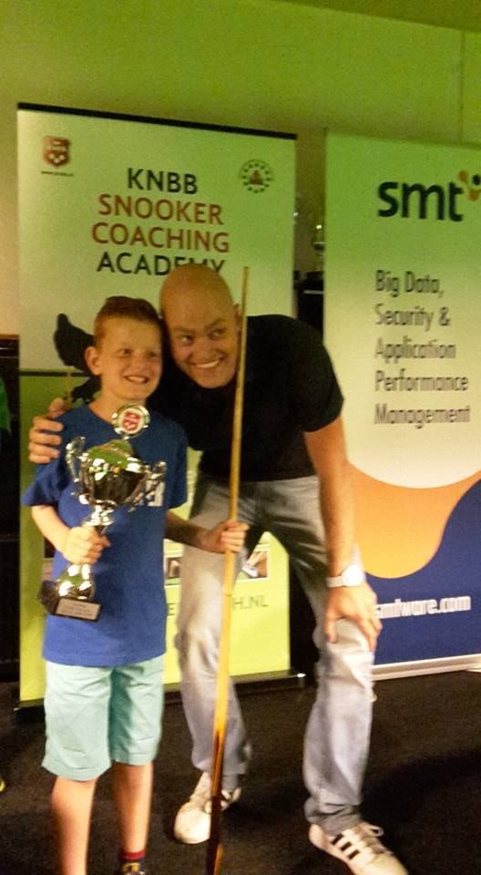 Een jongen met een trofee en een man met een snookerkeu lachen naar de camera terwijl ze voor de banners van het evenement staan.
