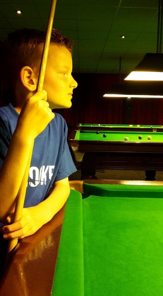 Een jongetje zit aan een pooltafel, met een keu in de hand, en kijkt aandachtig naar het spel. Geelachtig licht.