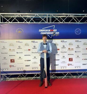 Jongeman in formele kleding met een keu in de hand tijdens het Europees kampioenschap snooker. Rode loper, podiumachtergrond.