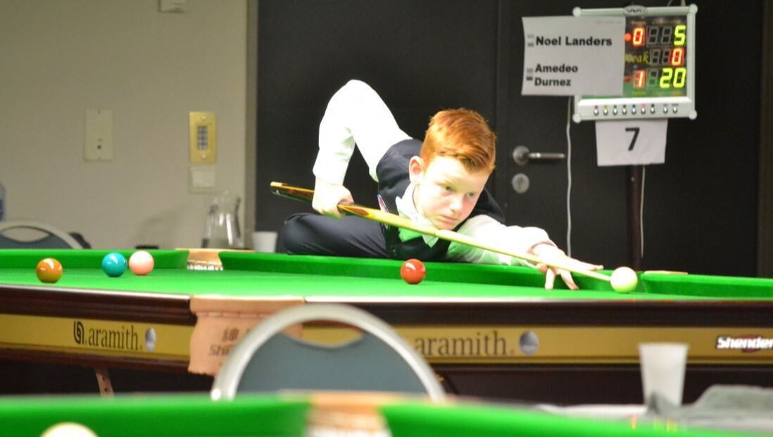 Jongen met rood haar speelt snooker, leunt over de tafel en mikt op een stoot, het scorebord op de achtergrond.