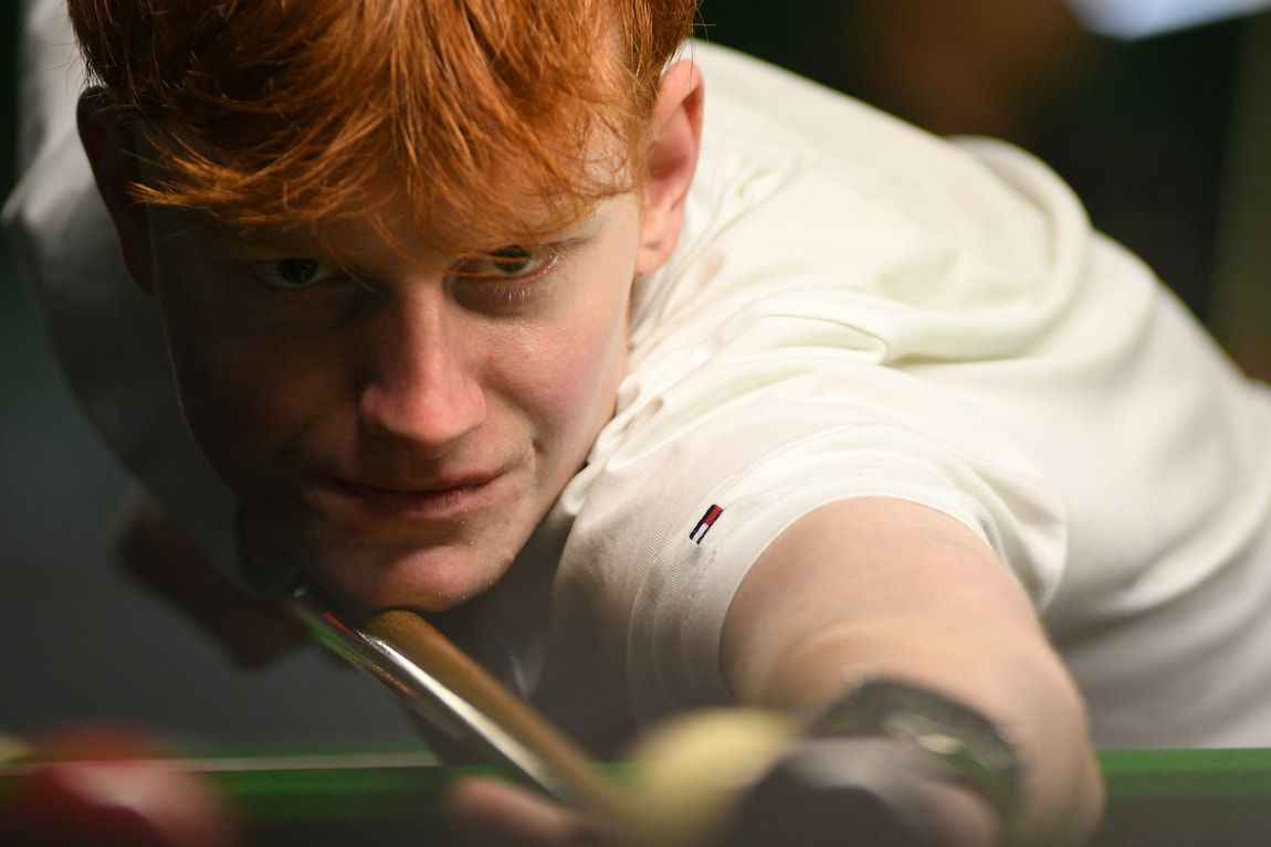 Man with red hair lining up a snooker shot, wearing a white shirt.