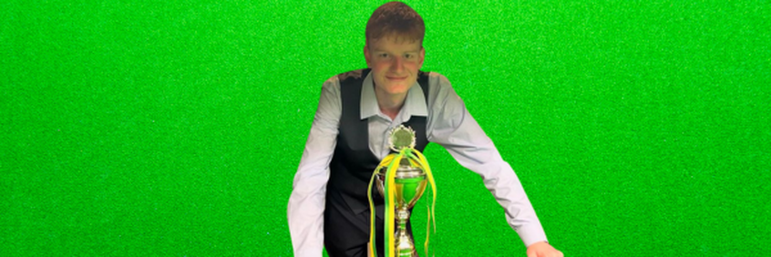 Persoon die een trofee vasthoudt tegen een groene achtergrond; draagt een vest en een lichtgekleurd shirt.