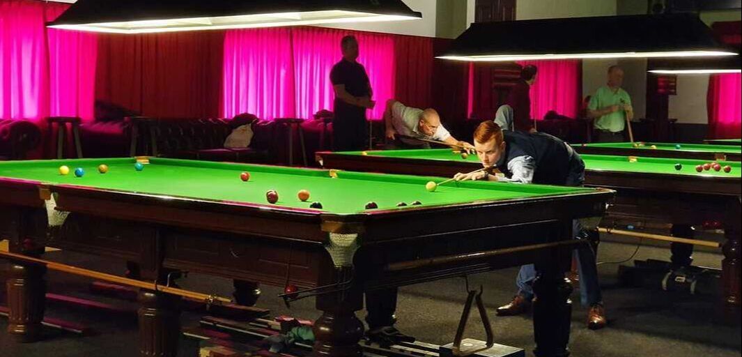 Mensen spelen snooker in een schemerige kamer met roze gordijnen en plafondlampen.