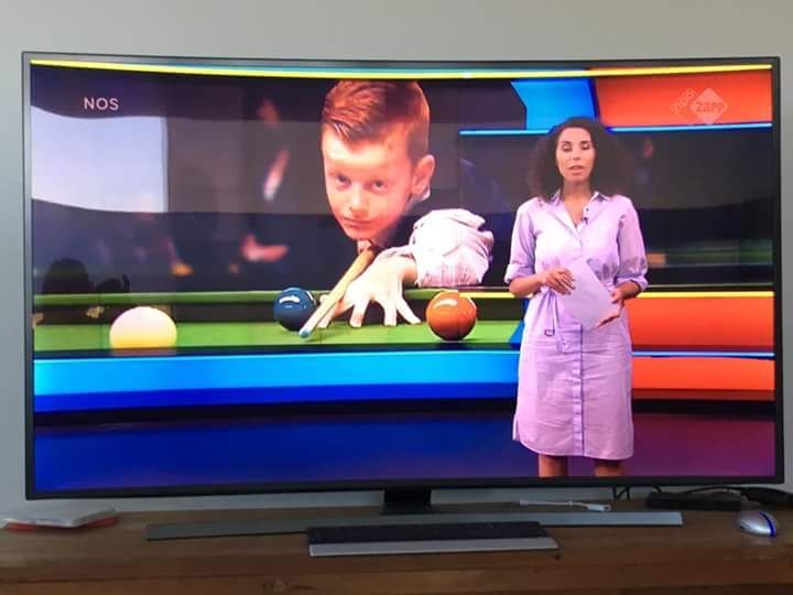 Een vrouw in een roze jurk presenteert het nieuws naast een jongen die op tv snooker speelt.