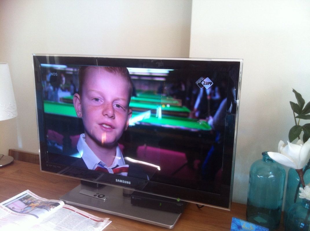 Jongen op tv-scherm met groene achtergrond; tv op houten bureau bij vaas en bloemen.