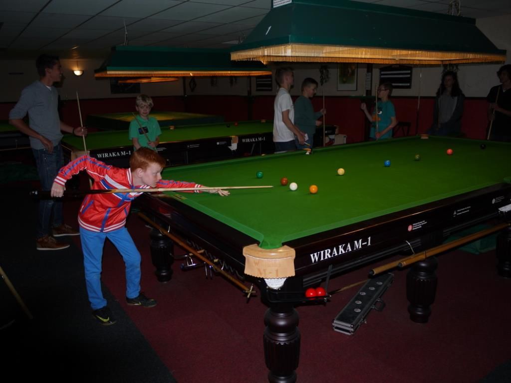 Jongen aan het poolen, binnen, meerdere mensen kijken toe. Groene tafel, kleurrijke kleding, gedempt licht.