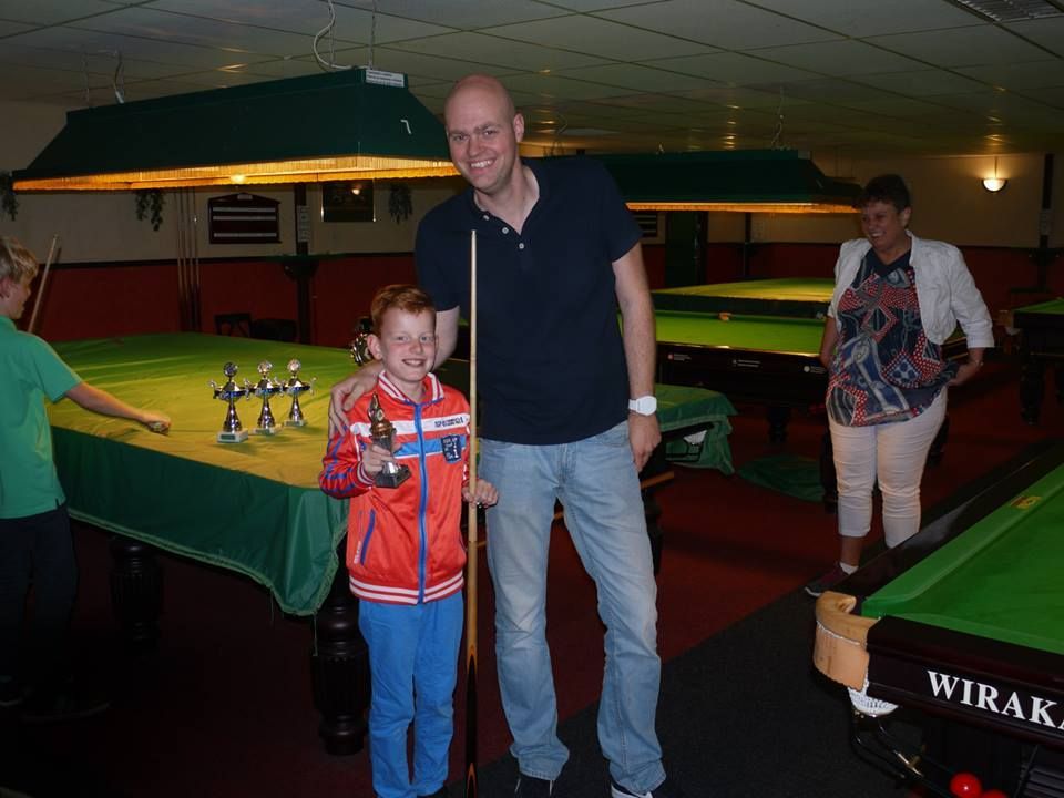 Man en jongen met trofee aan snookertafel, andere spelers en tafels op de achtergrond.