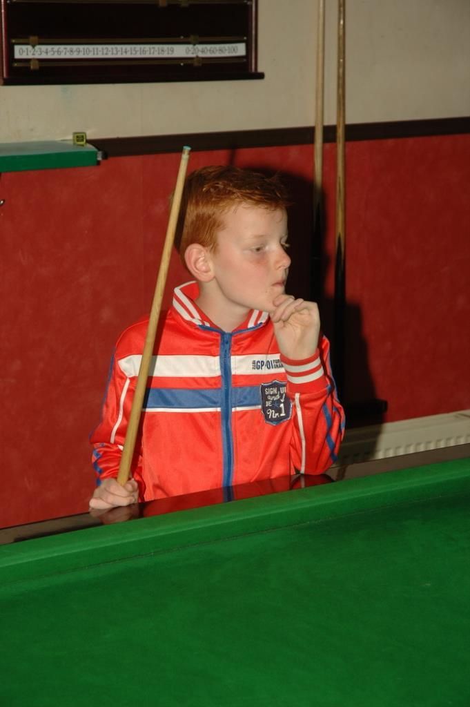 Jongeman met rood haar in een rood trainingspak overweegt een potje snooker op een groene tafel.
