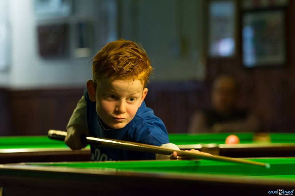 A young boy with red hair aiming a pool cue on a green table, concentrating, in a dimly lit room.